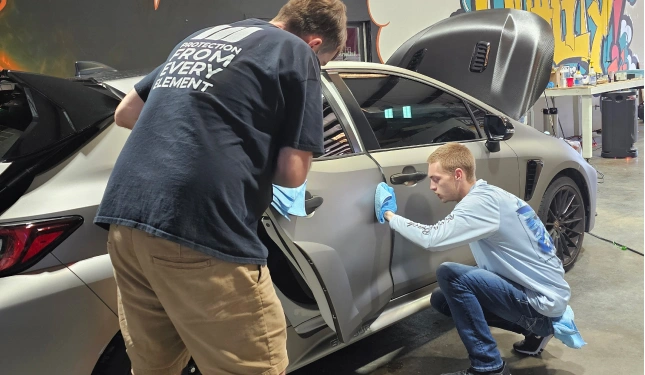 Two auto detailing technicians apply a protective film or vinyl wrap to the side of a matte gray car with open doors and hood inside a custom garage with graffiti-style wall art.