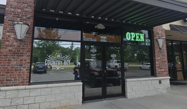 Exterior of a retail storefront labeled "Country Boy" with a logo featuring a cannabis leaf, arrows, and the words "Smoke • Vape • CBD • Delta," and a green neon "OPEN" sign in the window.