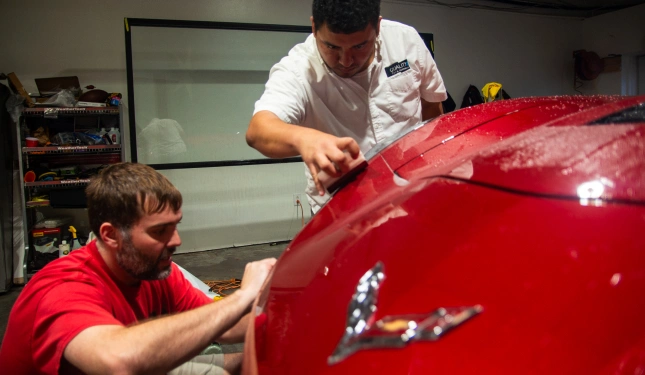 Two men carefully apply protective film to the hood of a red Corvette inside a detailing shop, with one person smoothing the surface and the other aligning the film.