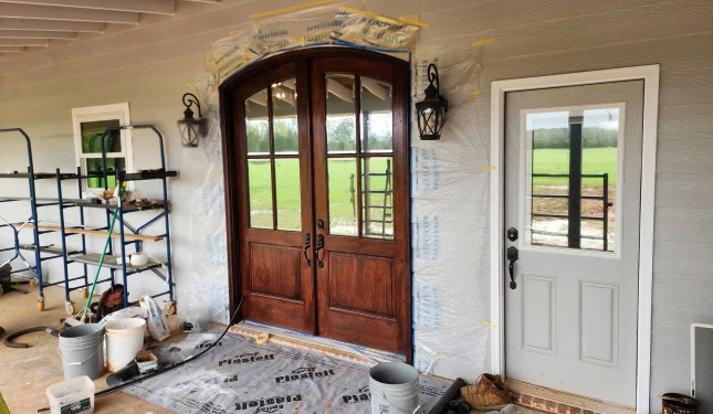 Wooden double doors with glass panes are prepped for staining or finishing, surrounded by taped-off plastic sheeting and work materials, next to a white door with a window on a covered porch.