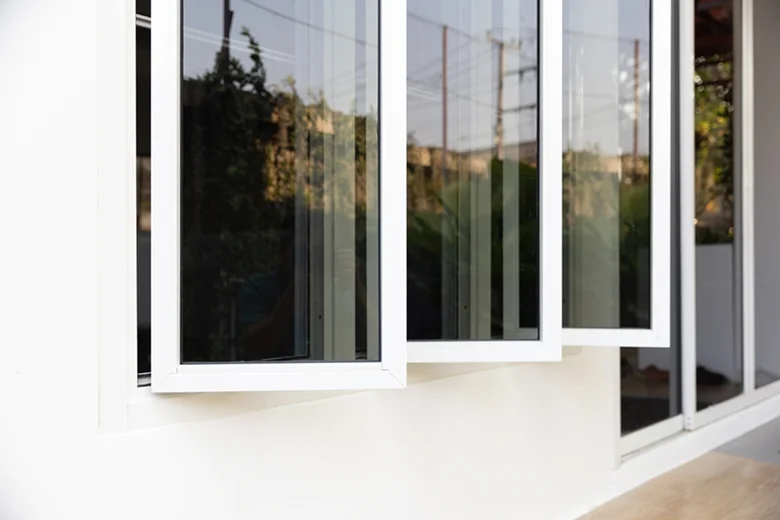 Three glass window panels with white frames partially open outward from a modern white wall, reflecting trees and outdoor structures.