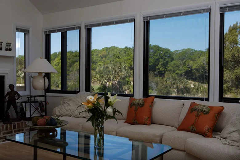 Living room with large black-trimmed windows showcasing a scenic view of lush green trees, featuring a beige sectional sofa with orange palm tree pillows and a glass coffee table with a flower vase.