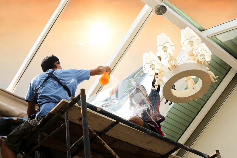 Man on scaffolding applying window film to a large, slanted skylight during sunset, with a chandelier nearby and an orange spray bottle in hand.