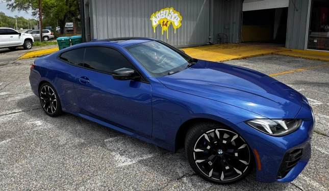 Sleek blue BMW coupe with blacked-out tinted windows and black multi-spoke rims parked in front of a building with a yellow "Wraptors" sign.