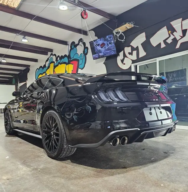 Shiny black Ford Mustang with tinted windows, quad exhaust tips, and black rims parked inside a detailing shop with colorful graffiti-style wall art and overhead lighting.