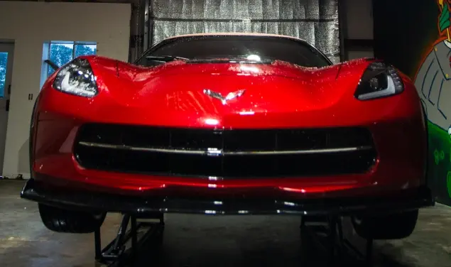 Front view of a red Chevrolet Corvette elevated on a lift in a garage, with headlights on and paint protection film being applied to the hood.