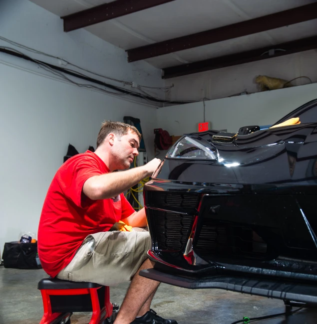 Man in a red shirt and khaki shorts sitting on a low stool carefully applying detailing work to the front of a glossy black sports car inside a garage.