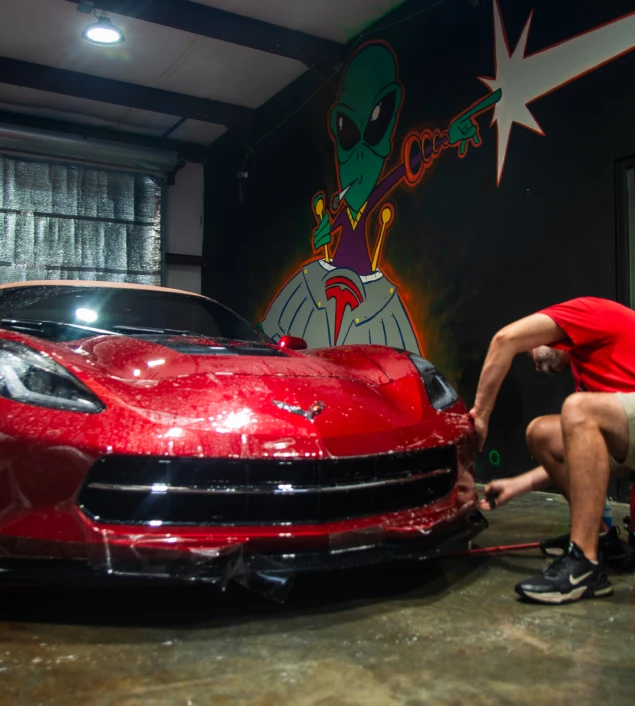 A red Corvette in a garage receiving a protective film installation on the front bumper, with a mural of a green alien holding a ray gun in the background.