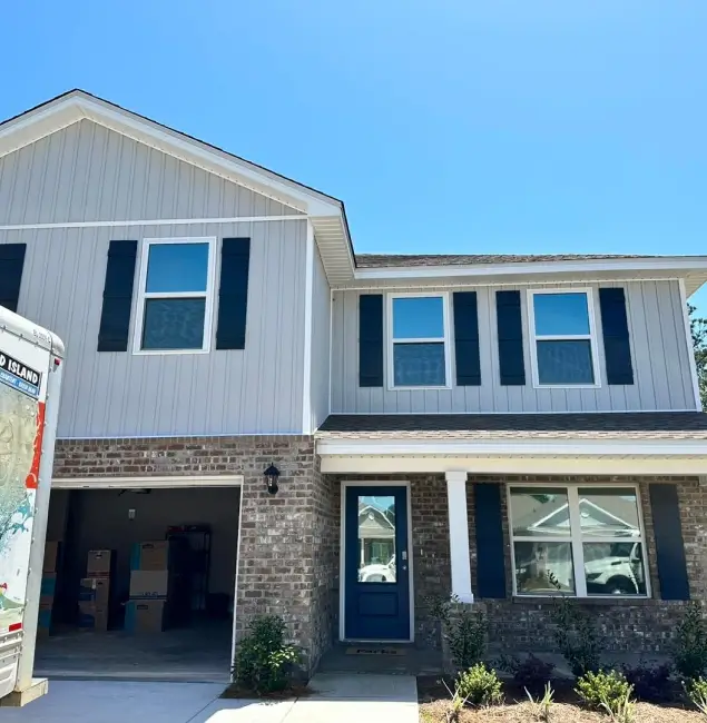 Two-story suburban home with a mix of light gray siding and brick exterior, black shutters, and a blue front door, with a moving truck and unpacked boxes visible in the open garage.