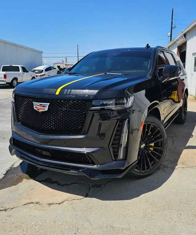 Black Cadillac Escalade SUV parked outside a garage, featuring a yellow racing stripe on the hood and matching yellow brake calipers.