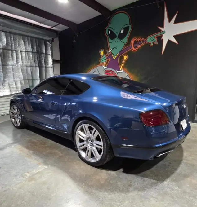Shiny blue Bentley coupe with dark tinted windows parked inside a detailing shop, in front of a mural of a green alien shooting a cartoon ray gun.