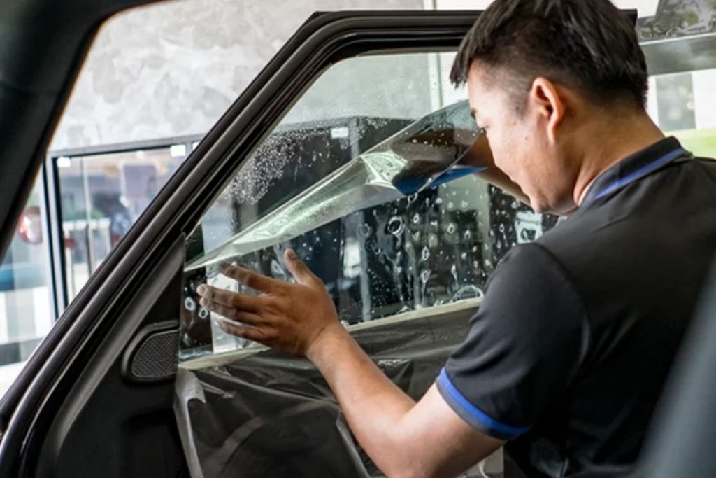 A professional installer carefully applies window tint film to a car window, with water and tools visible, illustrating the precision and care involved in quality tinting. The image reflects why Gulfport drivers choose window tinting for UV protection, privacy, and heat reduction.