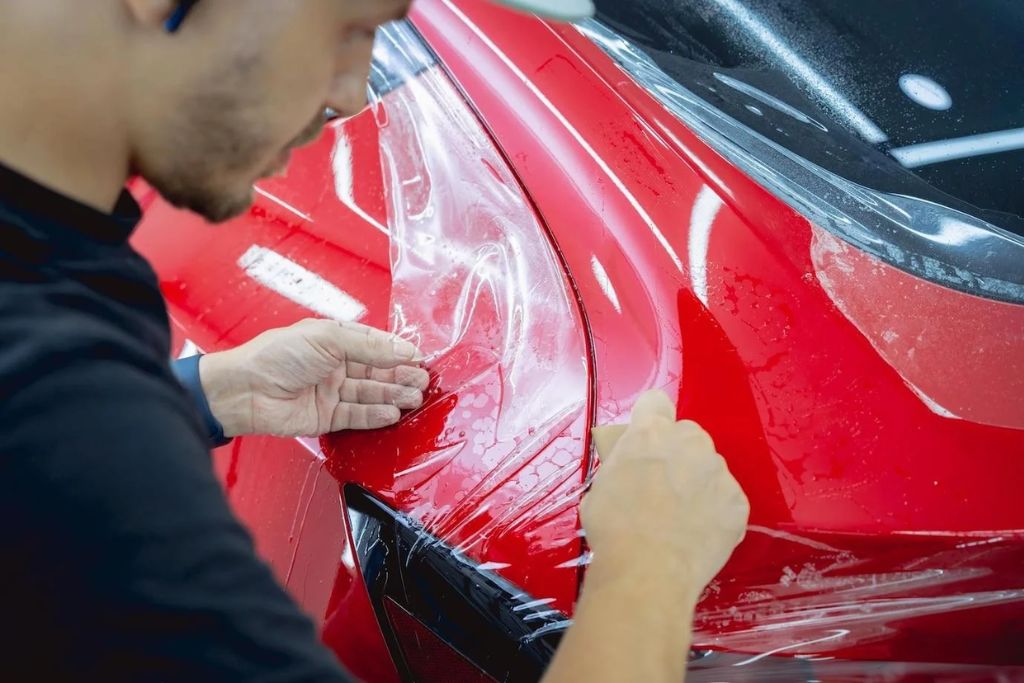 Close-up of a professional in Gulfport carefully applying Paint Protection Film to the front corner of a red car, showcasing the precision and benefits of this high-gloss, self-healing barrier in understanding what is Paint Protection Film.