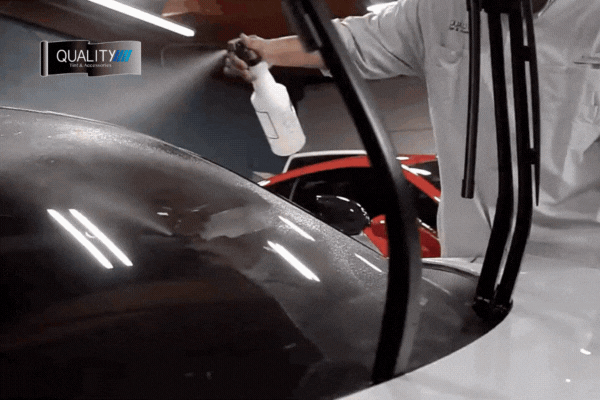 A technician from Quality Tint & Accessories sprays solution on a car’s rear windshield during a professional tint installation, with wiper blades lifted. The motion highlights precision, cleanliness, and attention to detail in the tinting process.