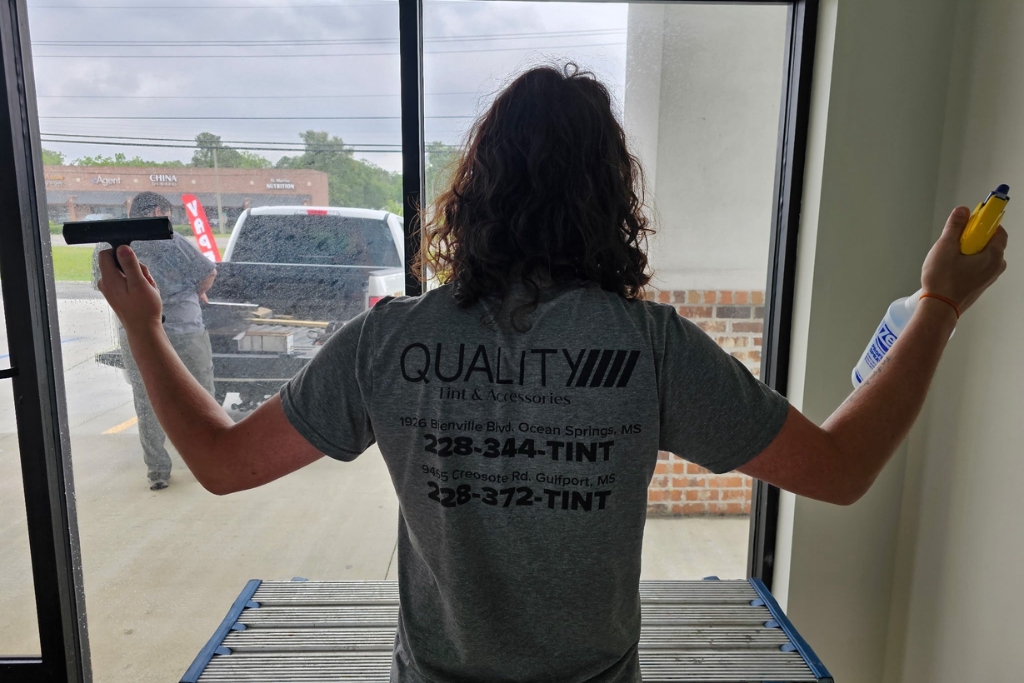 A professional installer from Quality Tint & Accessories stands in front of a large window, holding a squeegee in one hand and a spray bottle in the other, preparing to apply residential window tinting film. The worker wears a branded shirt displaying company contact information, emphasizing professional service.