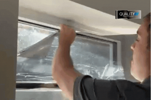 A technician from Quality Tint & Accessories applies residential window tinting to large angled windows inside a white brick room. One panel is mid-installation with dark film partially attached, while others are prepped for tint, reflecting both the interior lights and outdoor scene.