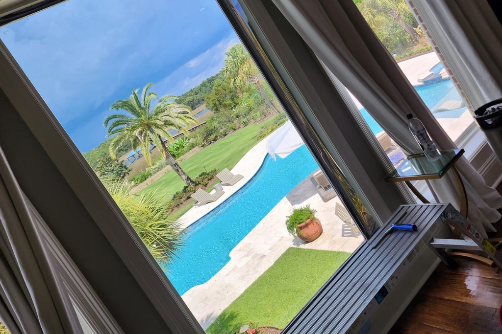A large floor-to-ceiling window overlooks a luxurious backyard pool and palm trees, with a residential window tinting film partially applied on one side. The film subtly darkens the glass, reducing glare while preserving the scenic view.