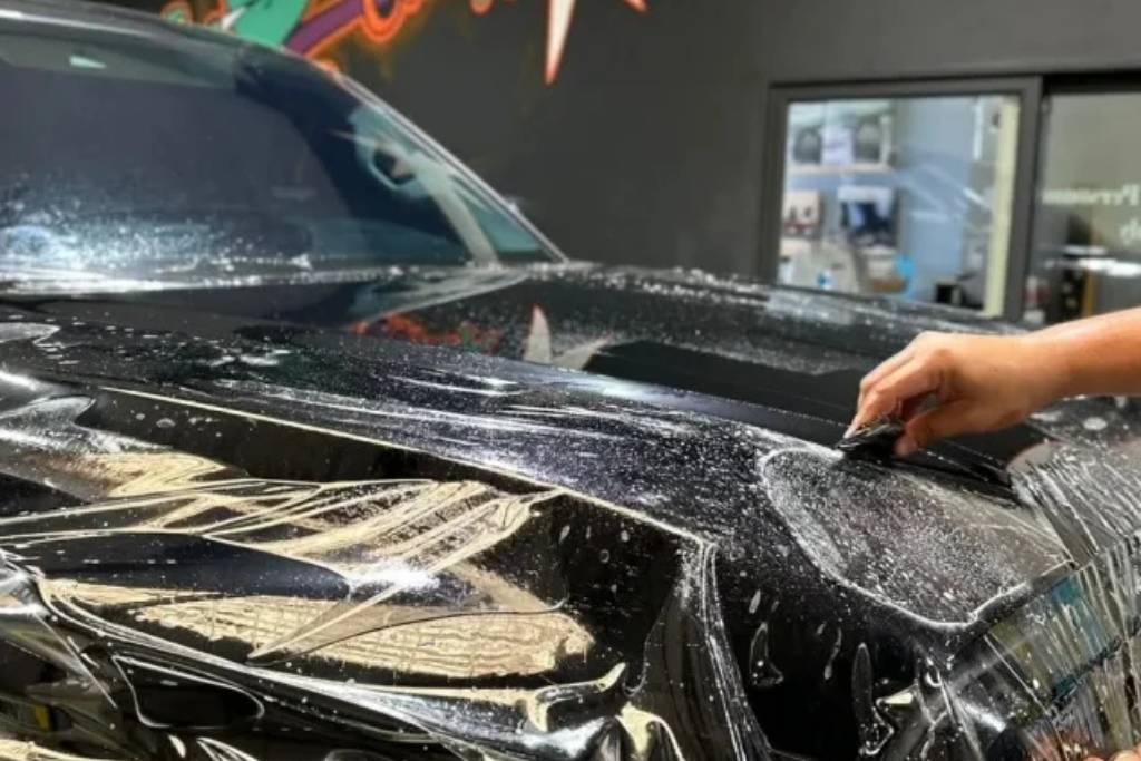 Close-up of a technician applying Paint Protection Film (PPF) on a black vehicle hood using a squeegee during the wet installation process.