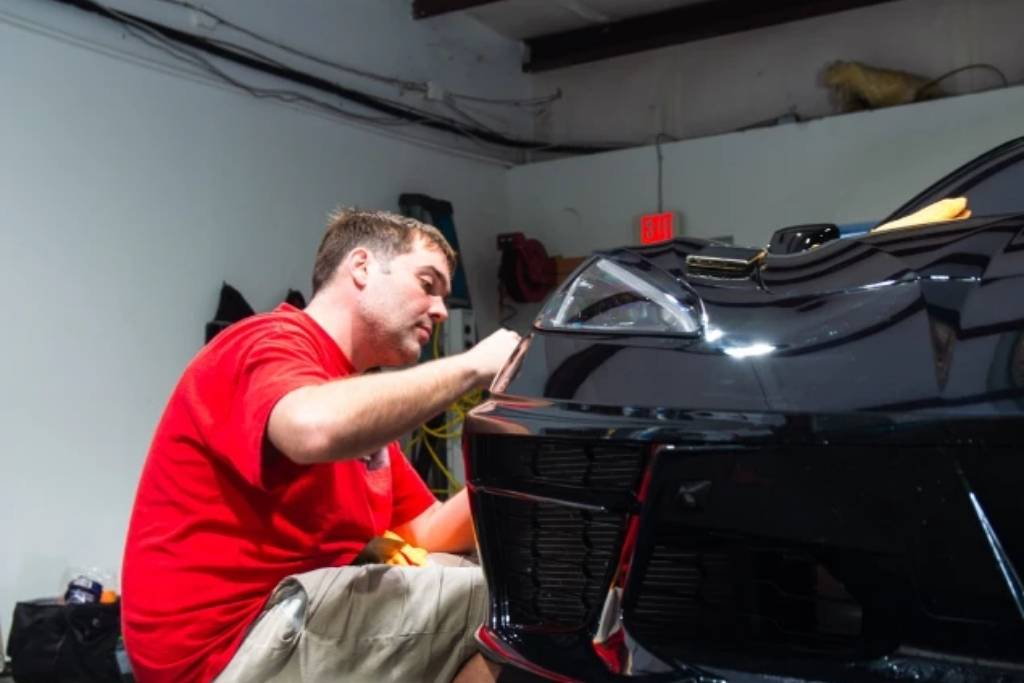 An auto detailing professional in a red shirt carefully applying paint protection to the front bumper of a black sports car inside a workshop.