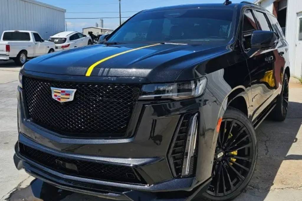 A black Cadillac SUV featuring custom detailing and a bold yellow racing stripe, parked outside a professional auto detailing shop.