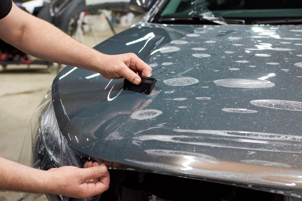 Technician applying paint protection film on a car hood using a squeegee to ensure smooth, bubble-free installation.