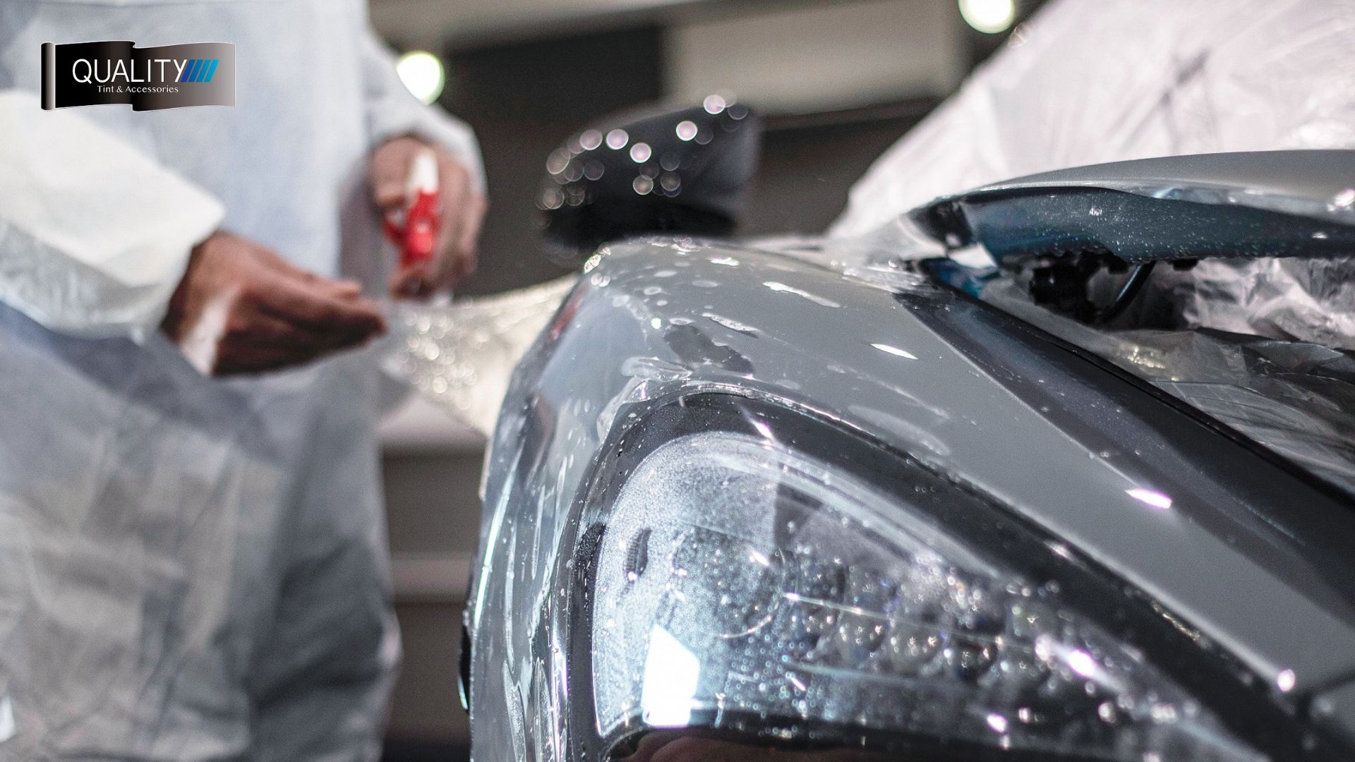 Technician in protective gear applies paint protection film to the front bumper and headlight of a glossy dark gray car.