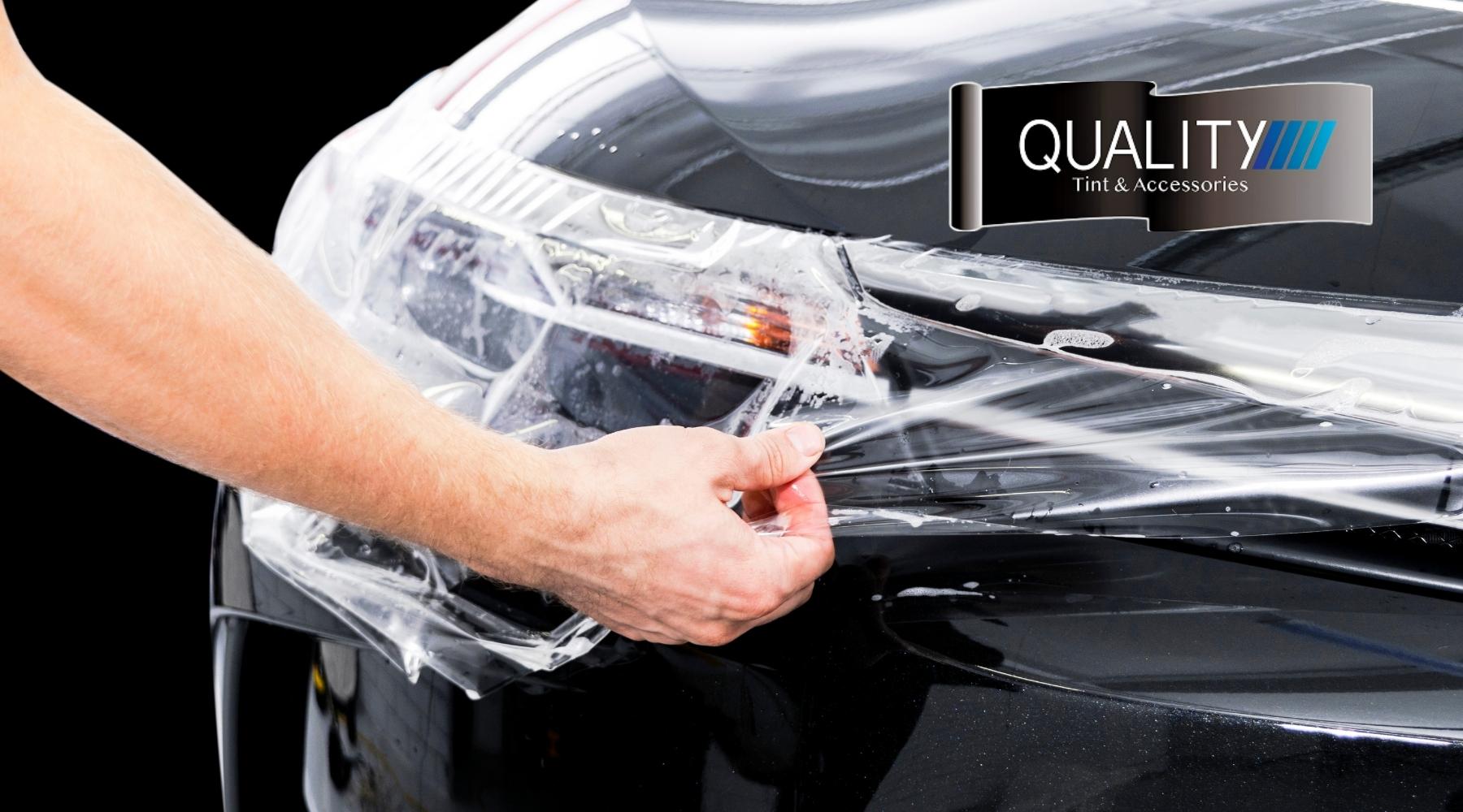 Technician applying transparent paint protection film to the front of a black vehicle with the Quality Tint & Accessories logo displayed.