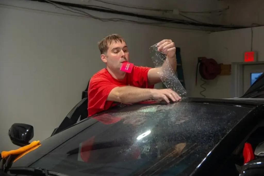 Technician in a red shirt applying window tint or paint protection film to the top of a car windshield inside a service garage.