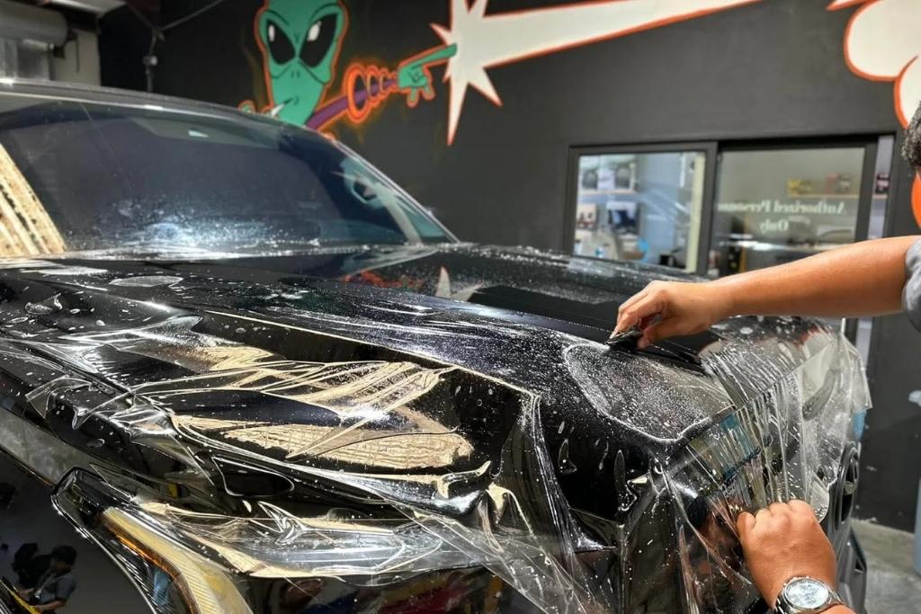 Technician applying paint protection film to the hood of a black SUV inside a shop with mural artwork in the background.