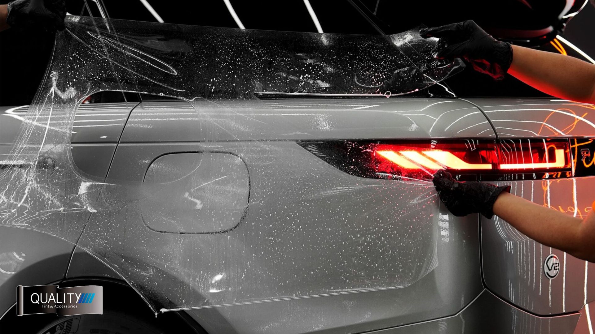 Two gloved hands stretch and align paint protection film over the rear side of a silver SUV, with red taillights illuminated and a cutout for the fuel cap visible.