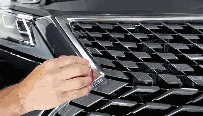 Detail shot of a person installing paint protection film on the chrome grille of a black SUV, pressing the film down with a pink tool.