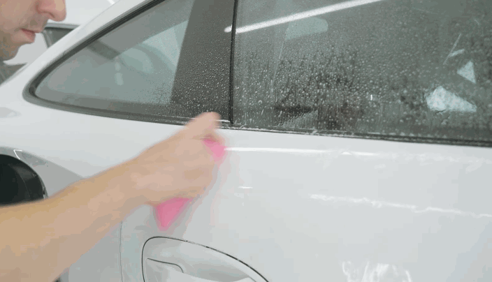 Technician smoothing out paint protection film on the rear door of a white car using a pink squeegee while the surface is still wet.