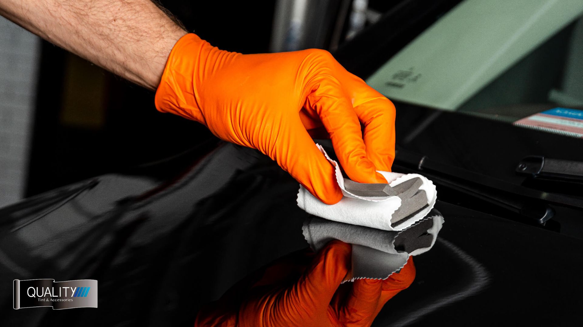 Close-up of a person wearing an orange glove applying ceramic coating to a black car surface using a folded microfiber cloth. The paint reflects the hand and cloth clearly.