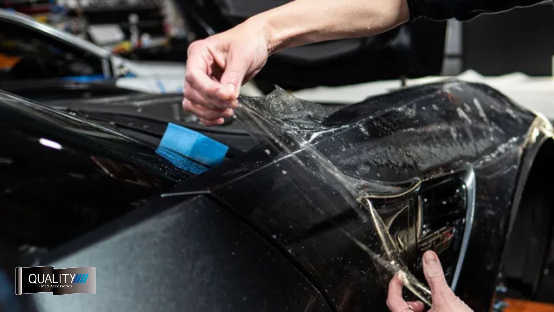 Close-up of a person applying paint protection film to the front fender of a black sports car, carefully smoothing it around the headlight area.