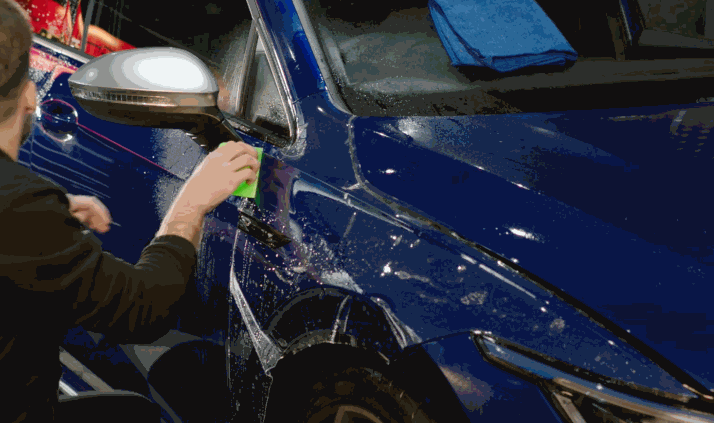 A man uses a green tool to secure paint protection film onto the front door and fender of a dark blue SUV, with visible water and reflections on the glossy surface.