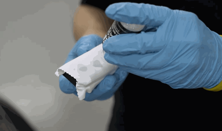 A person wearing blue gloves prepares a ceramic coating applicator by adding drops of product to a white microfiber-wrapped block.