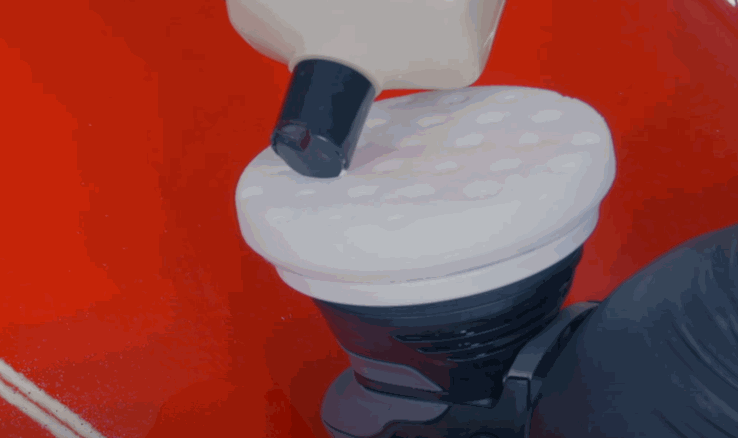 Liquid wax being dispensed onto a white foam pad attached to a car buffer over a red car surface. A gloved hand holds the buffer tool ready for application.