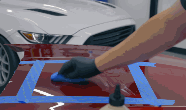 A gloved hand applies ceramic coating to a red car hood using a blue applicator pad within a taped-off section. A white sports car is visible in the background.